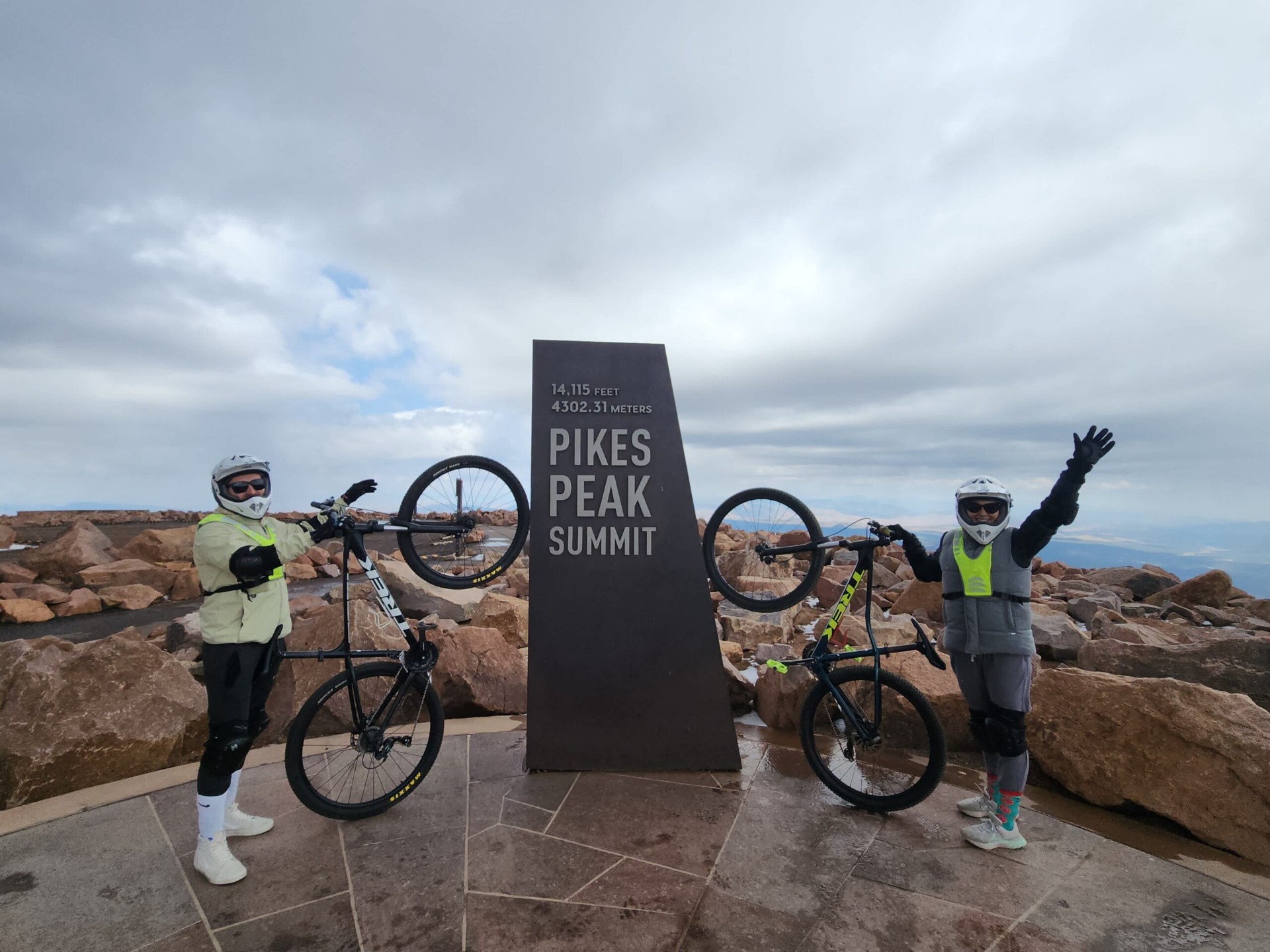 Two cyclists in helmets and pads celebrating with their mountain bikes at the 14,115 ft Pikes Peak Summit marker